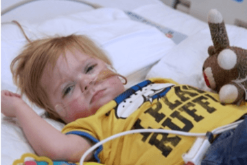 Young child lyign in hospital bed wearing yellow t-shirt and holding a stuffed toy