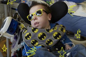 Photo of a young man in a blue shirt, wearing dark sunglasses with yellow rims reclining in a wheelchair with a batman printed neck pillow around his neck. He is in a hospital room and has a feeding tube attached to the wheelchair.  