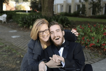 Woman in a grey sweater hugging a man in a tuxedo who is sitting in a wheelchair