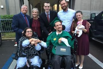 Photograph of a young woman and young man wearing a graduation cap and gown in wheelchairs holding hands with five individuals standing up behind them. One individual is holding an infant.