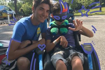 Photo of two young men in blue shirts sitting next to each other outdoors, one is in a wheelchair wearing a Mardi Gras mask and waving.