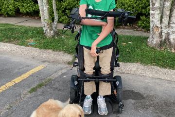 Bower wearing a green shirt and tan pants standing in his wheelchair smiling with his golden retriever everest