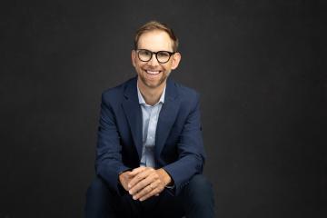 Michael Kutcher seated wearing a dark blazer with light shirt and dark rimmed glasses