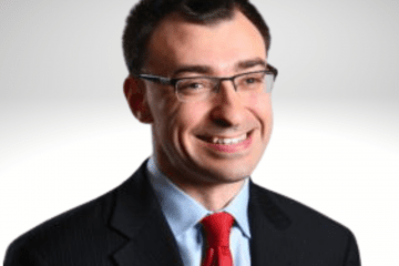 Jason has dark brown hair and is smiling wearing a blue shirt, red tie and a blazer.