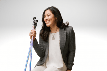 Xian Horn is sitting on a bench smiling, wearing white pants, white shirt and a brown blazer. She has walking sticks in her right hand. She has long brown hair and is smiling.