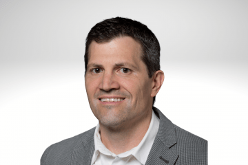 Christopher Modleski has dark hair and is smiling wearing a white button down and grey blazer