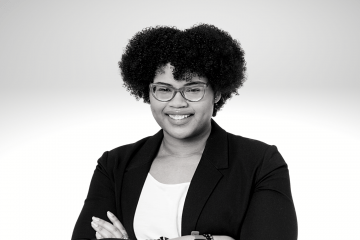Minelli Vasques has curly brown hair, she is smiling and wearing glasses. She has a white shirt and a black blazer with her arms crossed in front of her. 
