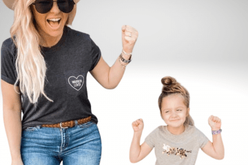 Fifi is a little girl standing in front of her purple walker wearing a grey t-shirt with her hands fisted in the air. She is smiling and has brown hair. She is next to her mom who is wearing a white cowboy hat, dark glasses, has long blonde hair and is smiling wearing a grey shirt and jeans.