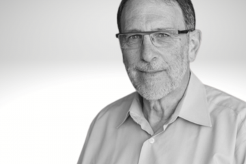 Peter Rosenbaum has dark hair, glasses and a short grey beard. He is wearing a white button down shirt.