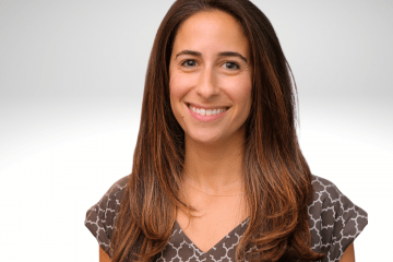 Jan has long brown hair and is pleasantly smiling for the camera. She is wearing a brown plaid dress with capped sleeves.