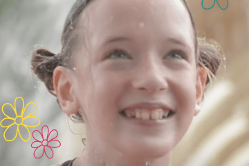 A little girl with blond pigtails and a big smile is in front of a fountain at a water park wearing her swim suit.