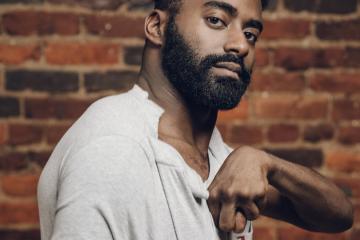 Jerron is standing in front of a brick wall wearing a white shirt with his left hand and arm up in front of his chest.