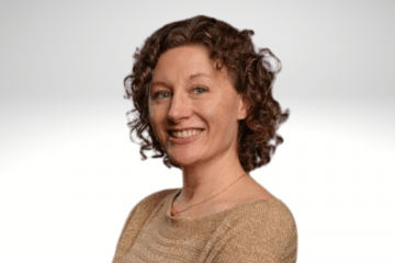 Dr. Heather Riordan has curley shoulder length light brown hair. She is wearing a tan short sleaved shirt and is smiling.