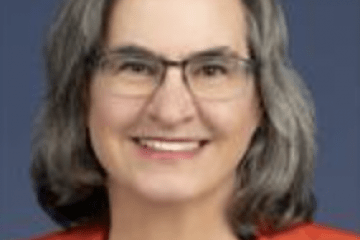 Dr. Kripke has shoulder length salt and pepper hair and is smiling wearing glasses and an orange blazer