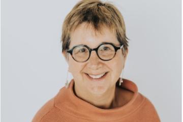 Ginny Paleg headshot. She has short brown hair and is wearing glasses and an orange turtleneck.